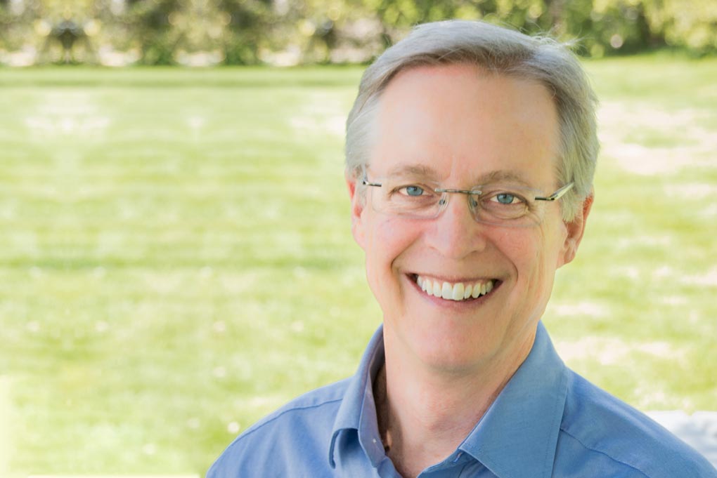 photograph of Dr. Leitner wearing a blue shirt standing in front of a green field and smiling